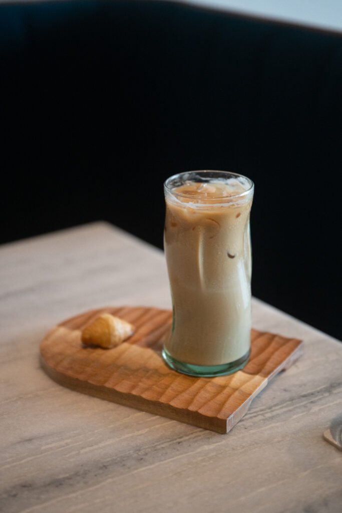  Decaf iced pistachio latte in a tall glass served on a wooden board with a mini croissant at Overflow Coffee, Golf City, Lucknow